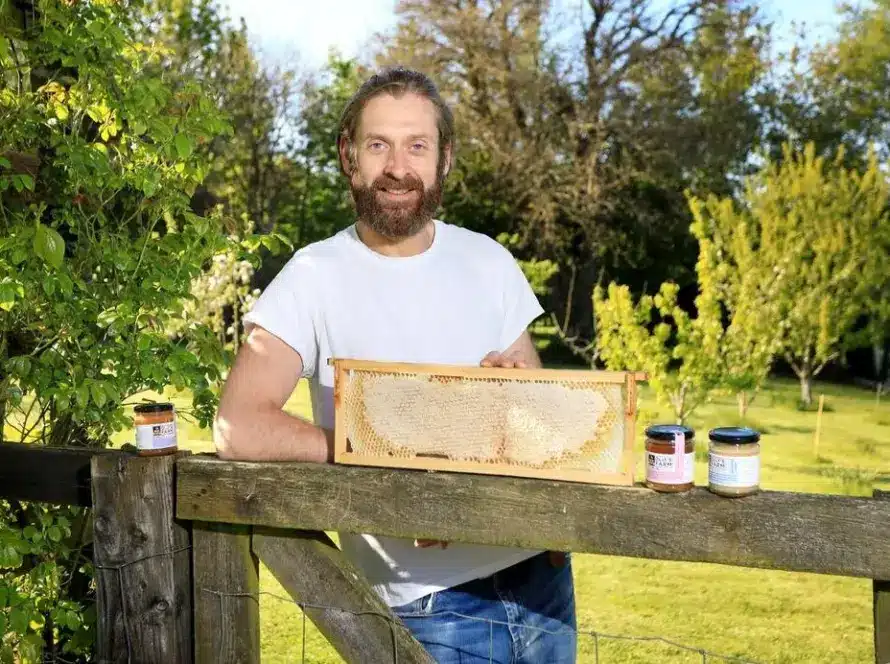 A man with a beard and a white t-shirt stands outdoors by a wooden fence, holding a honeycomb frame. Three jars of honey sit on the fence. Green trees and grass fill the sunny background.