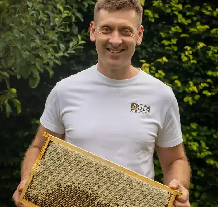 A smiling man in a white t-shirt stands outdoors, holding a wooden beehive frame full of honeycomb, with green foliage in the background.