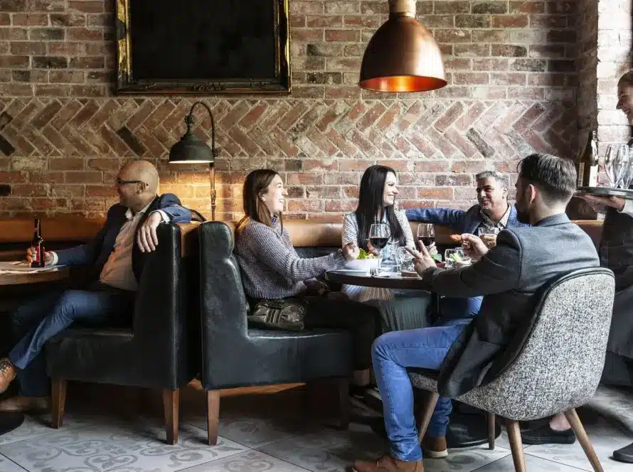Five people sit around a table in a stylish restaurant, talking and laughing. A server in an apron stands beside them holding a tray. The setting has exposed brick walls and warm lighting.