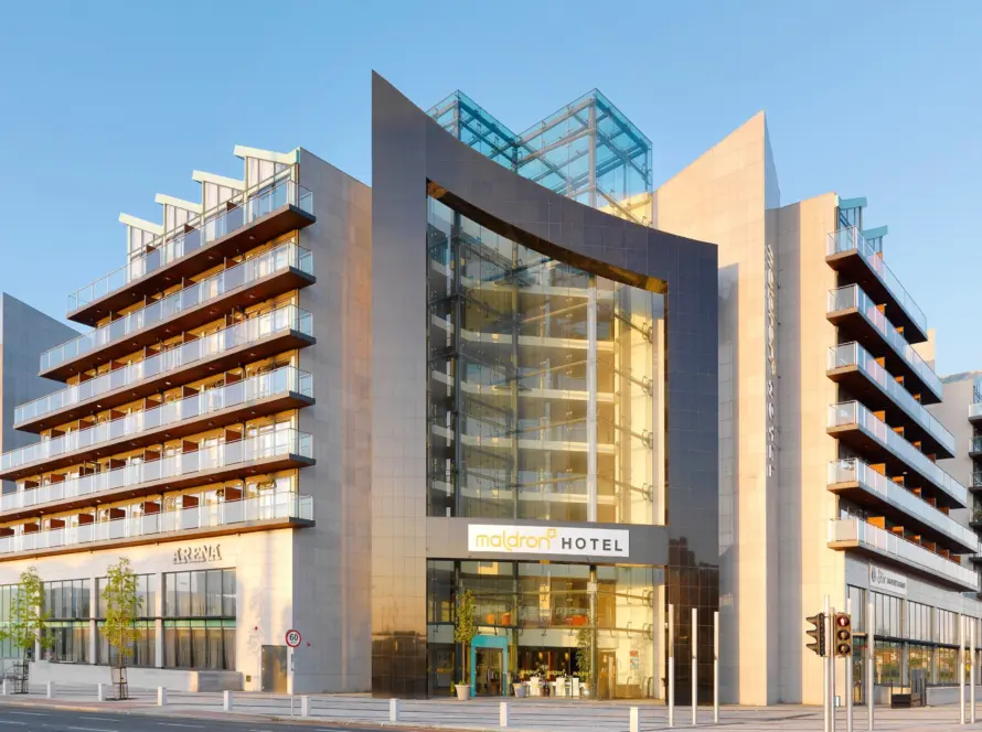 Modern multi-story hotel building with large glass entrance, angular architectural features, balconies, and signage reading "Maldron Hotel," captured under clear blue sky in daylight.