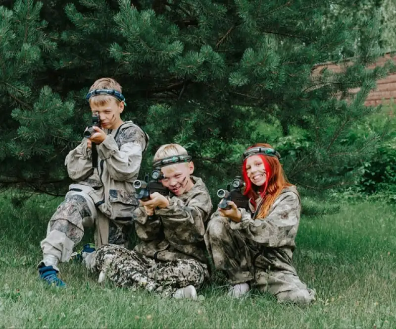 Three children in camouflage outfits and headbands holding toy laser guns crouch and kneel on green grass in front of trees, smiling and posing as if playing a team game outdoors.