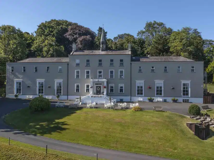 A large, symmetrical, gray manor house with white trim, surrounded by grass, trees, and outdoor seating areas on a sunny day. A curved driveway leads to the front steps and entrance.