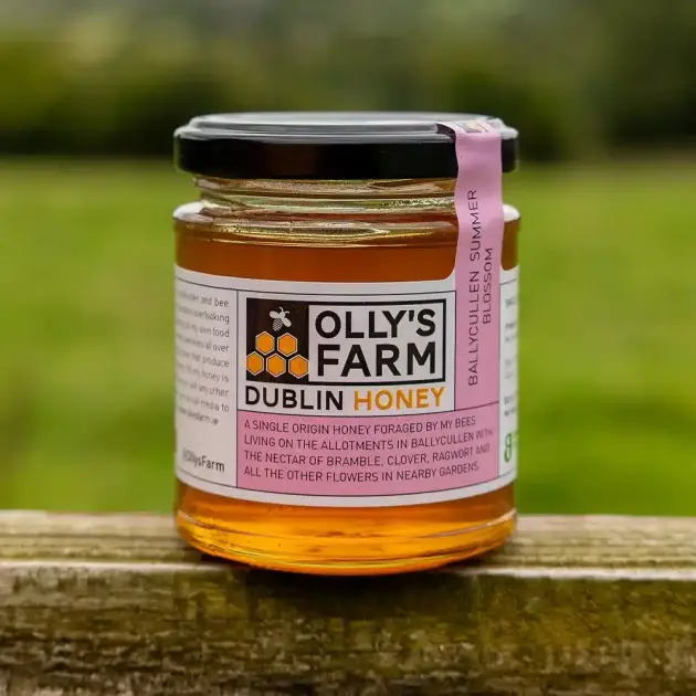 A jar of Ballycullen Summer Blossom Honey with a black lid rests on a wooden surface outdoors, set against a blurred green field. Its label details the honey’s origin and ingredients.