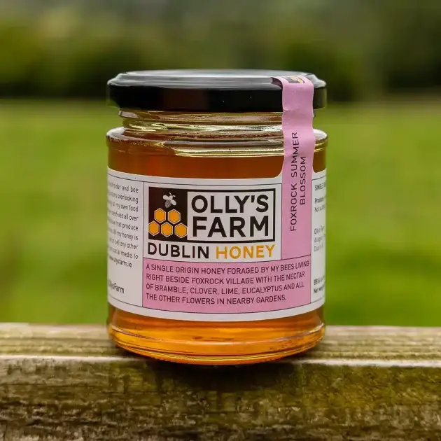 A jar of Foxrock Summer Blossom Honey with a black lid rests on a wooden surface outdoors. The label highlights it as single origin honey from Foxrock, made from a blend of local summer blossom flowers.