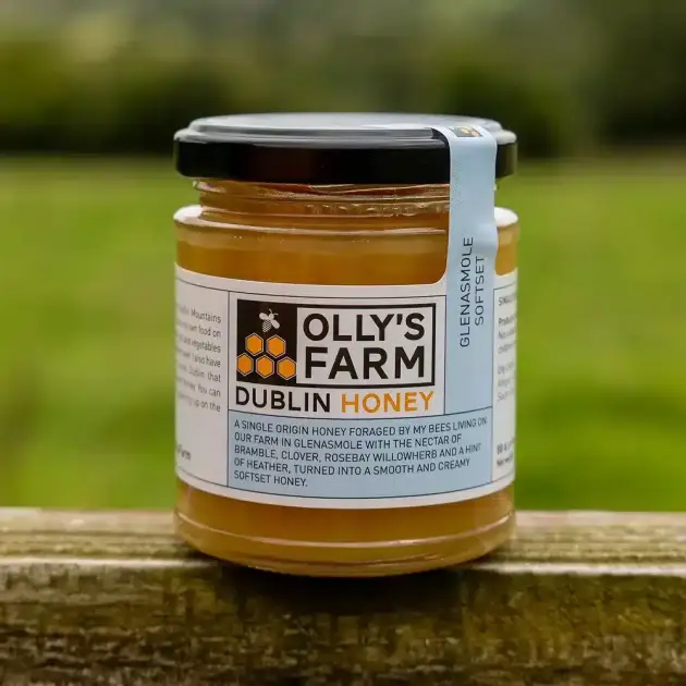 A jar of Glenasmole Softset Honey sits on a wooden surface outdoors, with a blurred green landscape in the background.