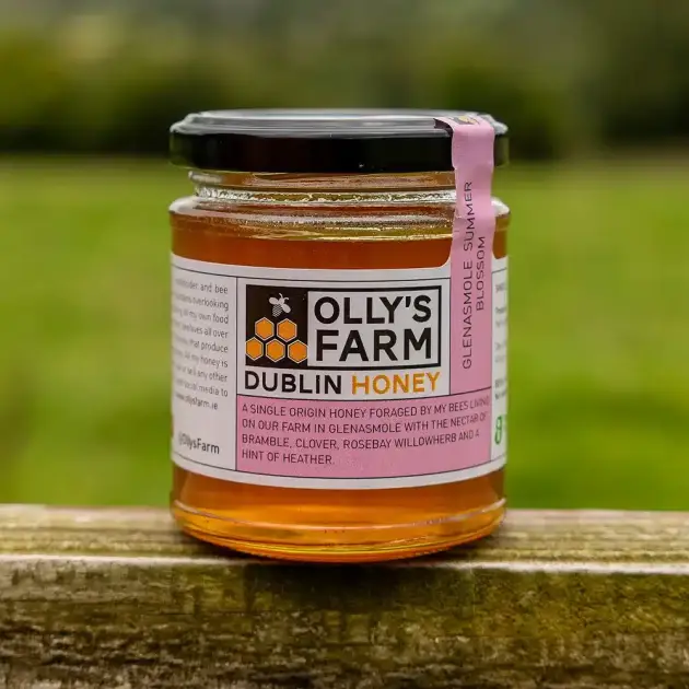 A glass jar of Glenasmole Summer Blossom Honey with a black lid rests on a wooden surface outdoors, its label highlighting floral sources. Blurred green grass appears in the background.