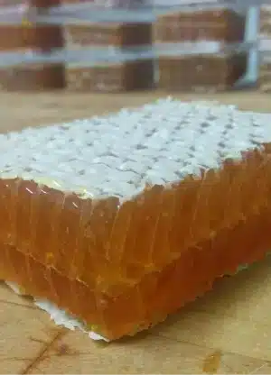 A close-up of a rectangular piece of honeycomb with golden honey inside, resting on a wooden surface. More honeycomb pieces are stacked in the blurred background.