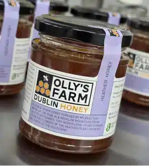 A close-up of a glass jar labeled "Olly’s Farm Dublin Honey" with a black lid and a purple ribbon. Other similar honey jars are blurred in the background on a reflective surface.