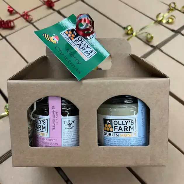 A cardboard gift box with two jars of Olly's Farm Dublin honey, labeled “Teeling Summer Whiskey” and “Dublin Honey,” with a green “Merry Christmas” tag, on a tiled surface with gold and red ribbon.