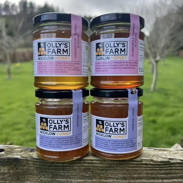 Four jars of Olly’s Farm honey are stacked on a wooden surface outdoors, with a grassy field and trees blurred in the background. Each jar has a different label and golden honey is visible inside.