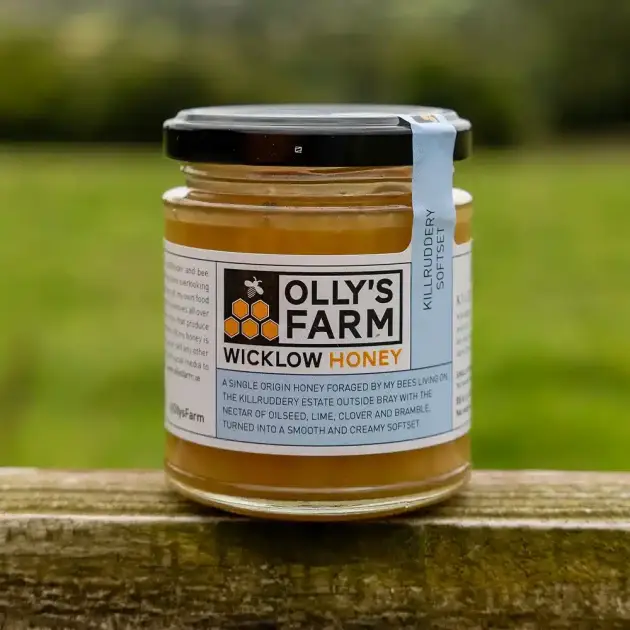 A jar of Killruddery Softset Honey rests on a wooden surface outdoors, with a blurred green field in the background and its label clearly visible.