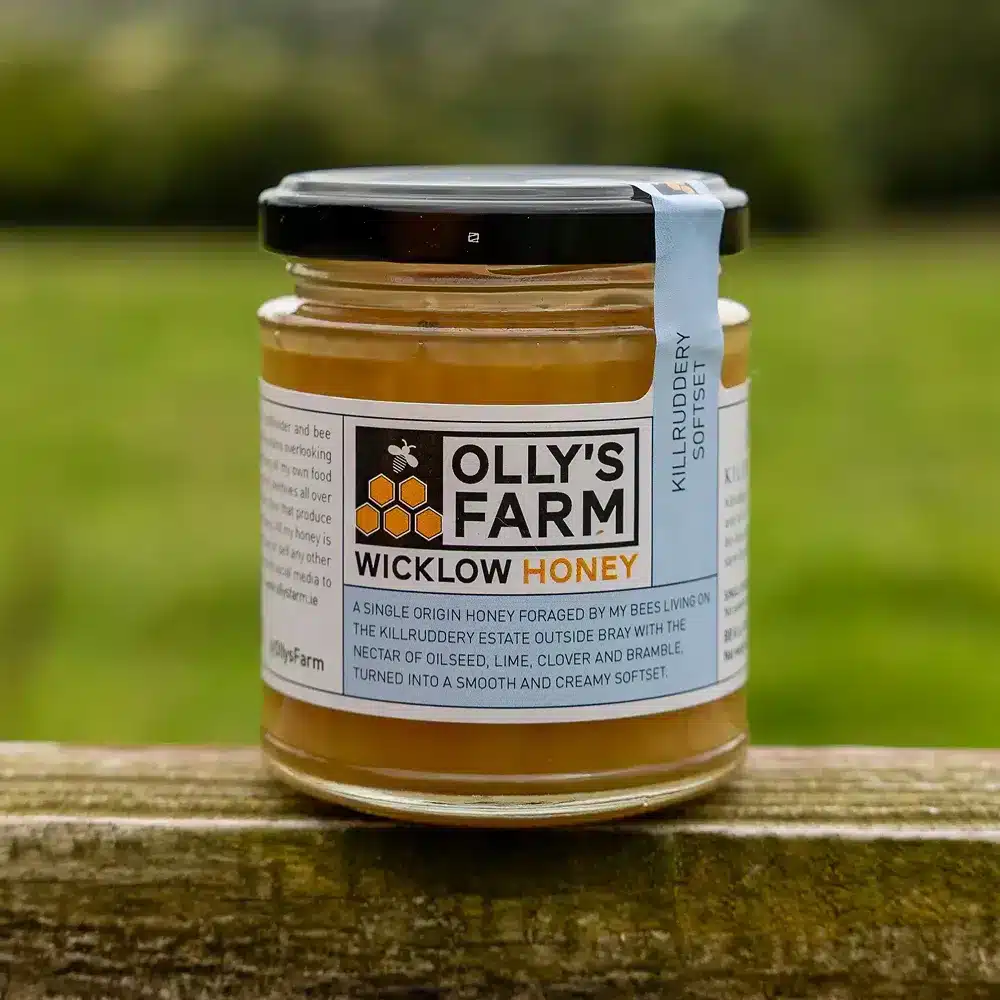 A jar of Killruddery Softset Honey rests on a wooden surface outdoors, with a blurred green field in the background and its label clearly visible.
