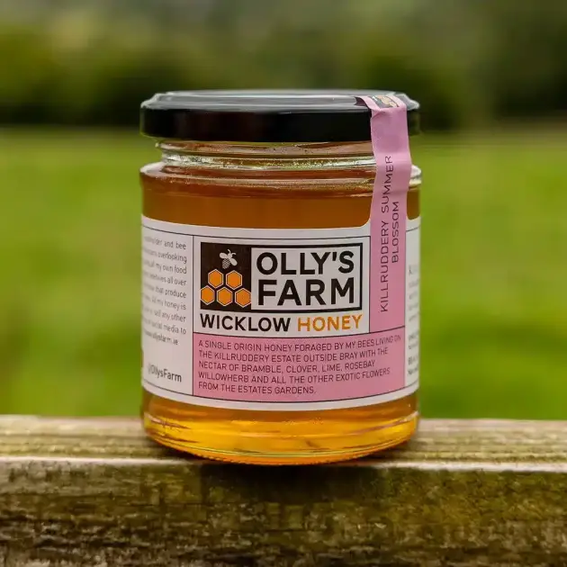 A glass jar of Killruddery Summer Blossom Honey with a black lid sits outdoors on wood, showing its label and golden honey inside, with a blurred background of green grass and trees.