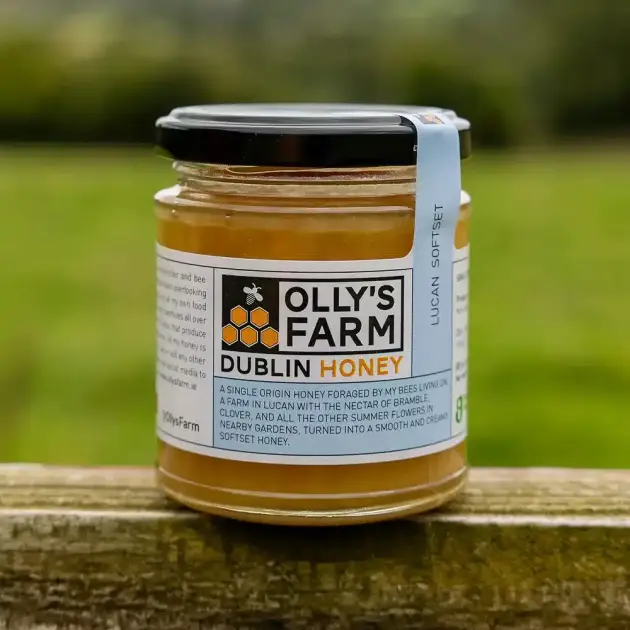 A jar of Lucan Softset Honey with a black lid and a white label showing a bee and honeycomb sits on a wooden surface outdoors, the green landscape in the background softly blurred.