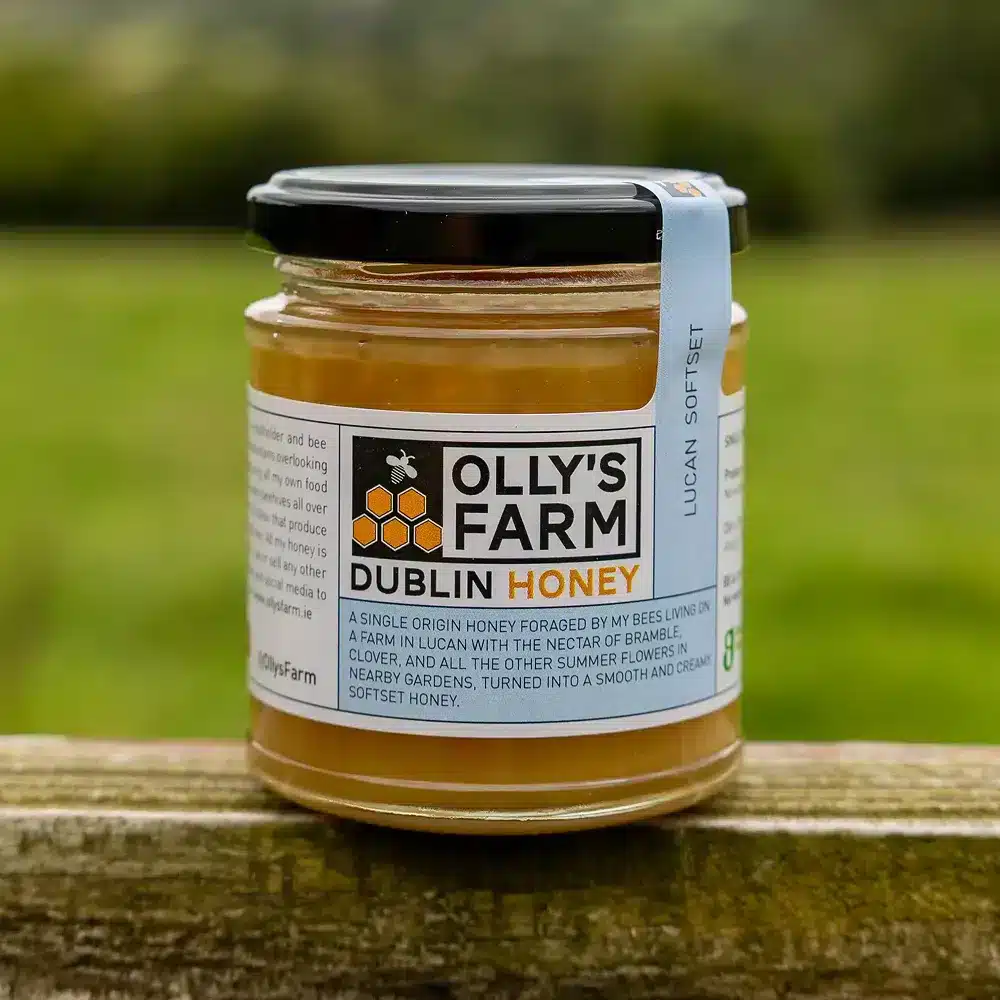 A jar of Lucan Softset Honey with a black lid and a white label showing a bee and honeycomb sits on a wooden surface outdoors, the green landscape in the background softly blurred.