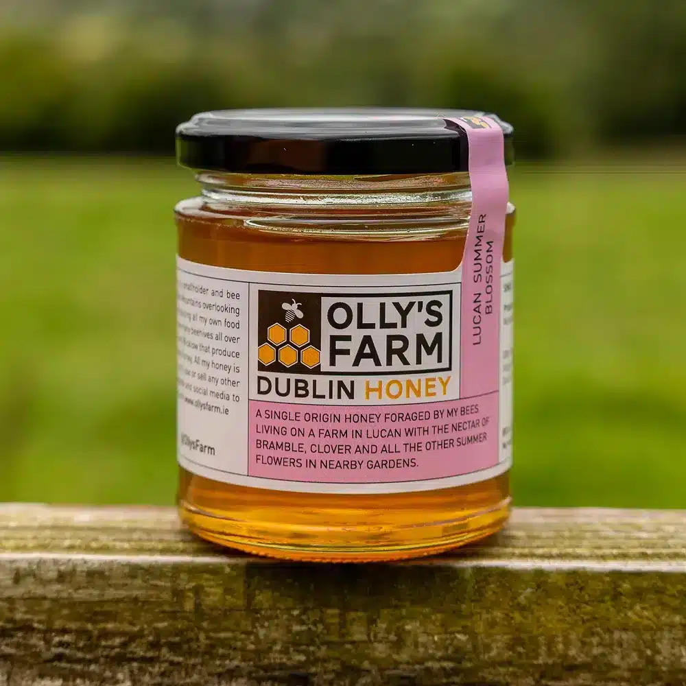 A jar of Lucan Summer Blossom Honey sits on a wooden surface outdoors, with a grassy field in the blurred background. The label highlights it as single origin honey made from local summer flowers.