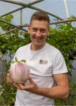 A smiling man in a white shirt stands in a greenhouse, holding a large, round, light purple eggplant with green leaves. Lush green plants fill the background.