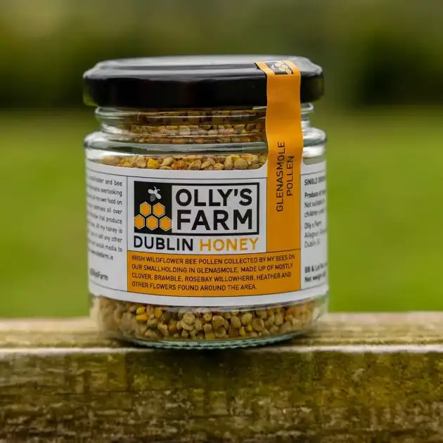 A glass jar labeled "Glenasmole Pollen," filled with bee pollen, sits on a wooden surface outdoors. The label notes it's locally collected by bees in Glenasmole, Dublin.