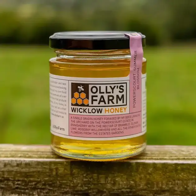 A clear jar of Powerscourt Summer Blossom Honey with a black lid sits on a wooden surface outdoors. The label features text and a pink ribbon marked “Powerscourt Summer Blossom,” displaying the honey’s golden color.