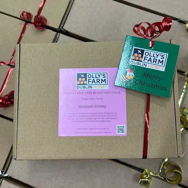 A brown gift box labeled "Olly's Farm Twin Blossom Honey Gift Box" is tied with red ribbon and a green "Merry Christmas" tag, with similar boxes stacked in the background.