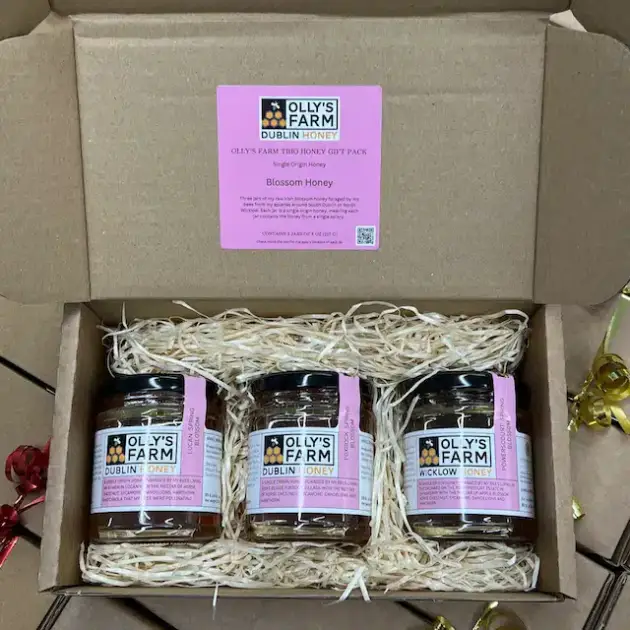 The Olly's Farm Trio Blossom Honey Gift Box includes three pink-labeled honey jars nestled in straw, with a "Olly’s Farm Dublin Honey" card inside the lid and colorful ribbon at the edge.