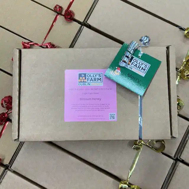 A brown Olly's Farm Trio Blossom Honey Gift Box with a pink label, festive green tag, and silver ribbon sits among similar boxes and gold and red ribbons.