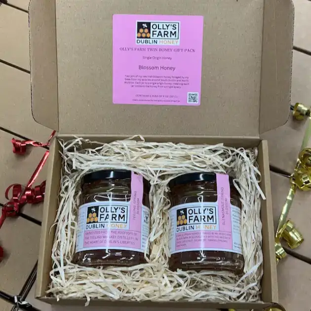 An open Olly's Farm Twin Blossom Honey Gift Box sits on a wooden surface, holding two jars of Dublin honey nestled in shredded paper. The box lid has a pink "Blossom Honey" label, with colorful ribbons lying nearby.