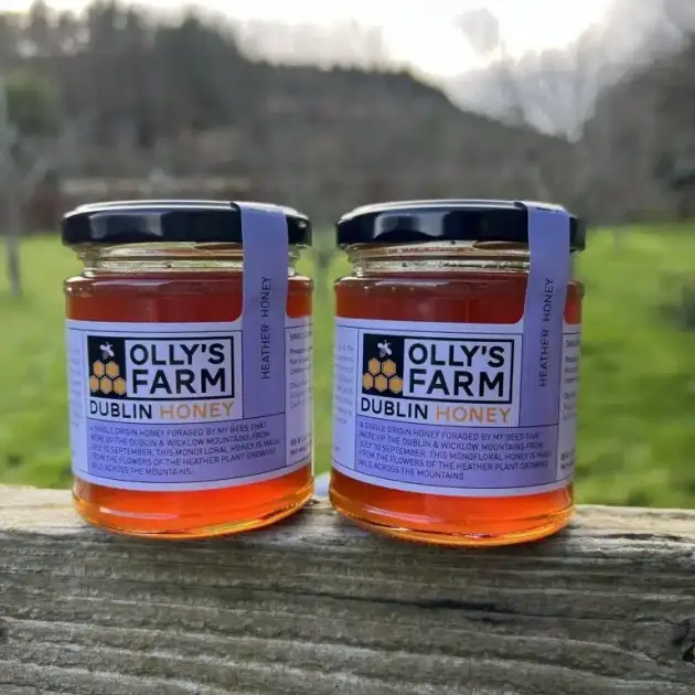 Two jars of amber heather honey from the "Subscription - 2 jars of heather honey every 3 months" sit on a wooden fence outdoors, featuring black lids and purple labels with a grassy field and trees blurred in the background.