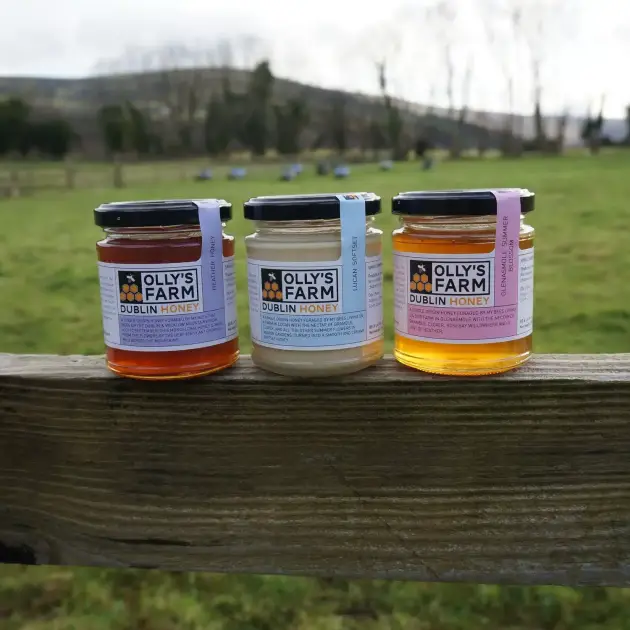 Three jars of Subscription - Heather, Softset & Blossom honey sit on a wooden fence, each with a different colored label and varying honey shades, against a backdrop of a grassy field and trees.
