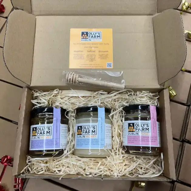 The Olly's Farm Trio Honey Gift Box includes three jars of honey, a wooden dipper, and an info card, all arranged on shredded paper in a cardboard box.
