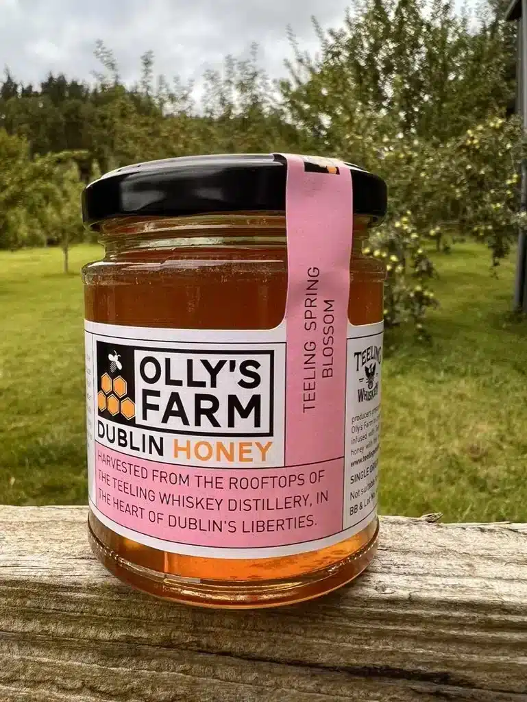 A jar of Teeling Spring Blossom Honey rests on a wooden ledge outdoors, with an orchard behind it. The label notes it’s harvested from rooftop hives at the Teeling Whiskey Distillery.