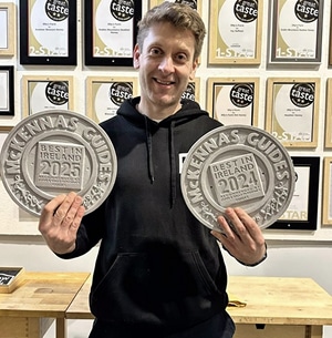 A smiling man in a black hoodie holds two "McKennas Guides Best in Ireland" awards for 2024 and 2025. Framed certificates cover the wall behind him.