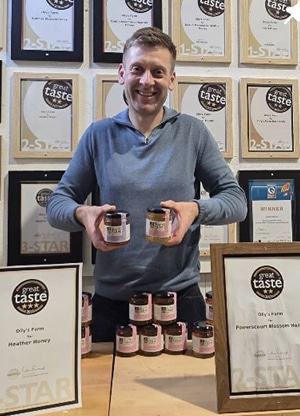 A smiling man in a blue hoodie holds jars of honey in front of a display of awards and certificates. Several jars of honey are arranged on the table, and framed "Great Taste" awards are visible.