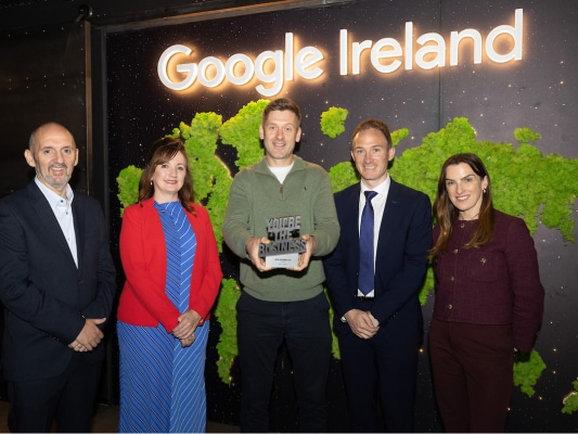 Five people stand in front of a green moss wall with a glowing "Google Ireland" sign. The person in the center holds a trophy that reads "Your Business." All are smiling and dressed in business or smart casual attire.