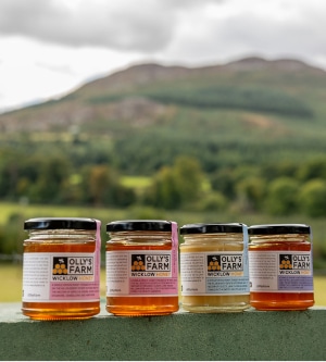 Four jars of Olly’s Farm Wicklow honey are displayed on a ledge outdoors, with a scenic, blurred background of green fields, trees, and hills under a cloudy sky.