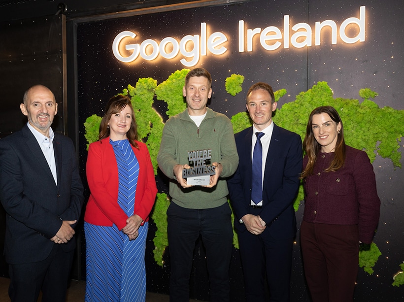 Five people stand smiling in front of a "Google Ireland" sign; the person in the center holds an award plaque that reads "You're the Business." The background features a green moss design shaped like a world map.