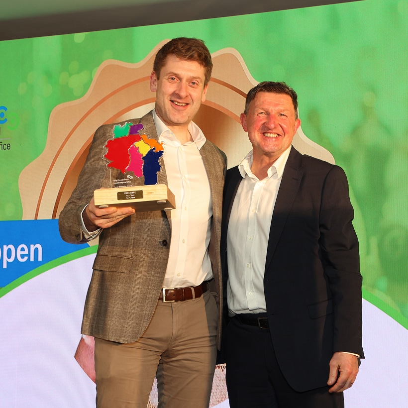 Two men in suits stand smiling; one holds a colorful trophy shaped like a cluster of regions. They are in front of a bright, green-themed backdrop at an awards event.