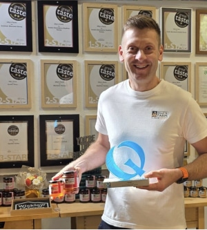 A man in a white shirt stands smiling, holding an award and jars of preserves. Behind him is a display wall filled with framed "Great Taste" award certificates and a table with jars and gift-wrapped items.