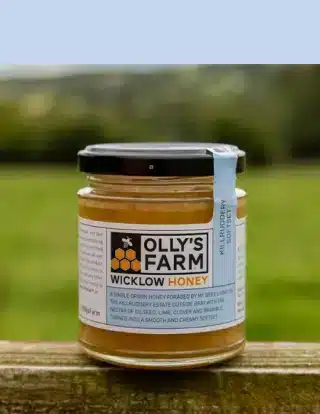 A jar of Olly's Farm Wicklow Honey with a blue seal sits on a wooden surface outdoors, with a blurred grassy background.