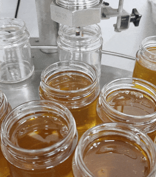 Honey is being poured into clear glass jars on an automated filling machine in a factory setting. The jars are arranged in rows and move under the dispenser as honey fills each container.