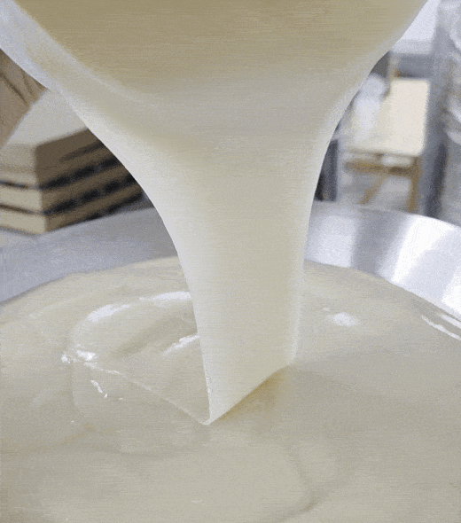 Thick, creamy white liquid is being poured smoothly from above onto a flat surface, creating glossy swirls as it spreads.