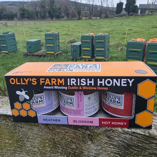 A box of the Heather, Blossom & Hot Honey Trio sits on a wooden fence, with green fields and beehives in the background.