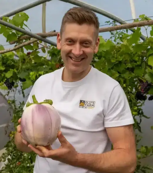 A smiling man in a white shirt stands in a greenhouse, holding a large, round, pale purple squash or eggplant. Green vines and clusters of grapes hang in the background.