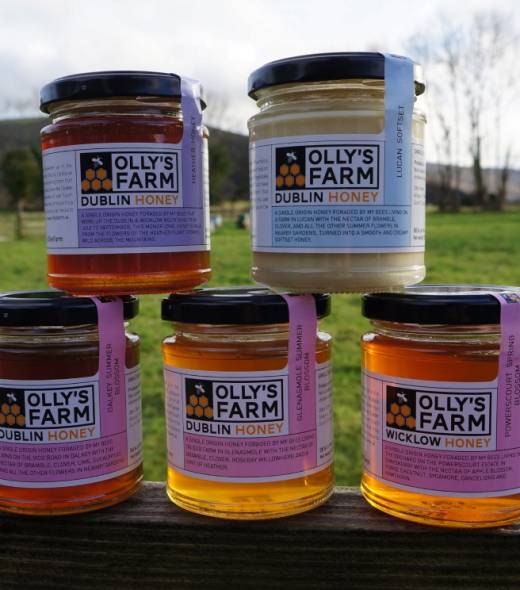 Five jars of Olly’s Farm honey, each labeled with different varieties, are arranged on a wooden surface outdoors, with a grassy field and trees visible in the background.