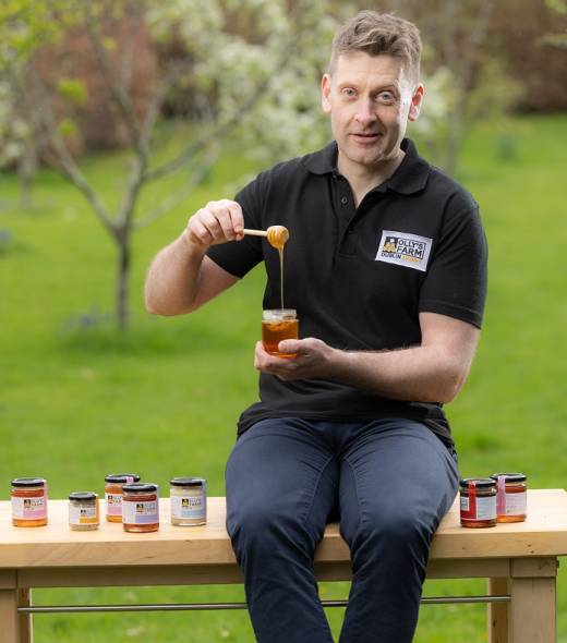 A man in a black polo shirt sits on a wooden bench outdoors, holding a jar of honey and dipping a honey dipper into it. Several jars of honey are displayed on the bench beside him, with a green garden in the background.